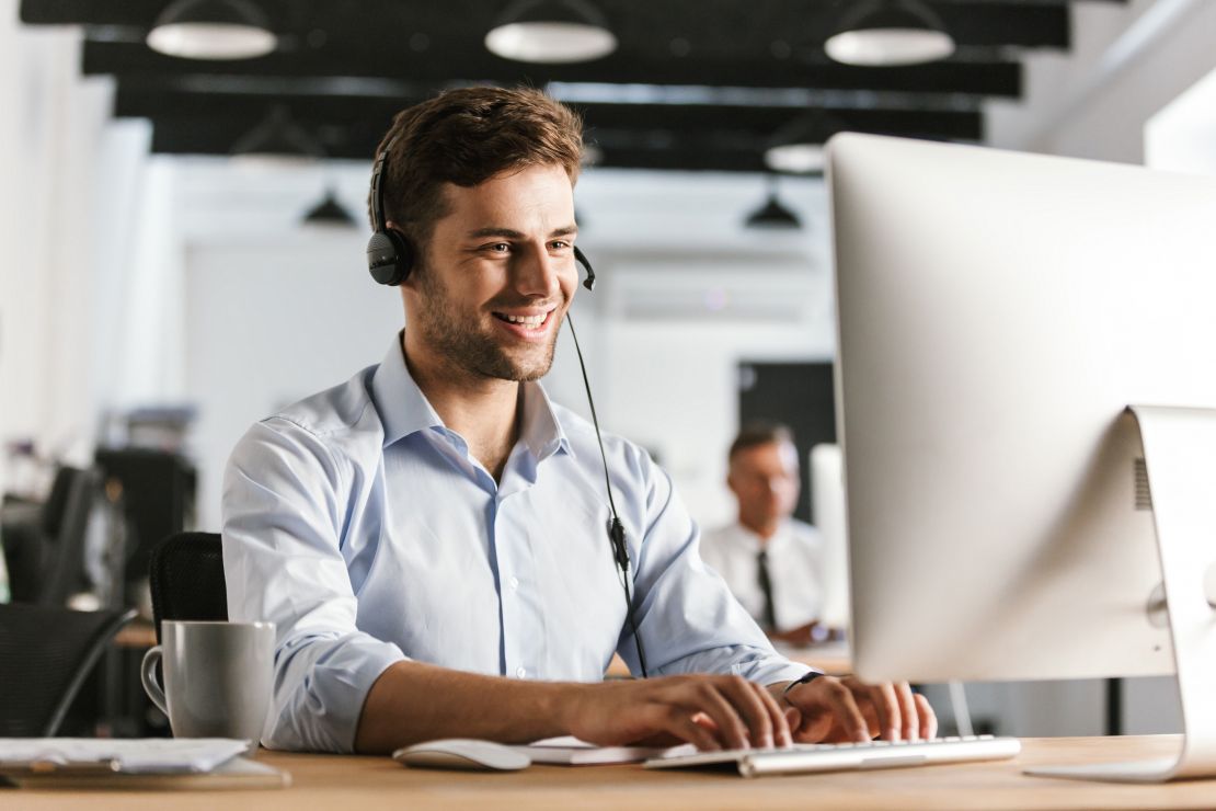 Junger Mann in hellem Hemd mit Headset am Schreibtisch