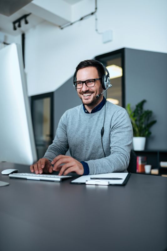 Mann im grauen Pullover mit Headset am Schreibtisch